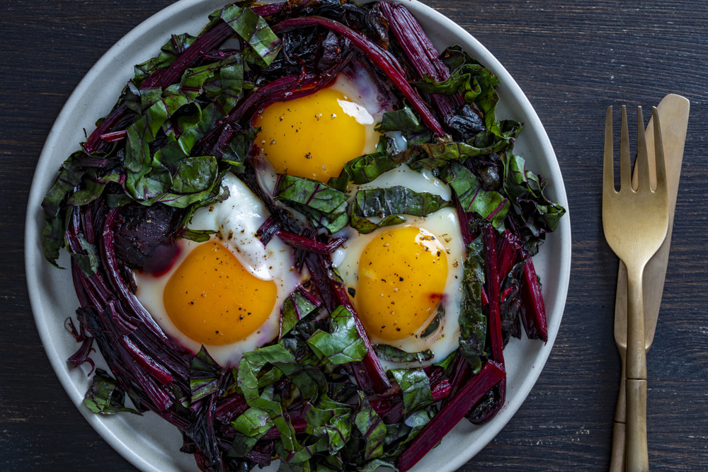 A nutritious breakfast: three fried eggs placed on sauteed beet greens and stems on a white plate with gold cutlery. High-protein vegetarian keto diet concept. A healthy breakfast