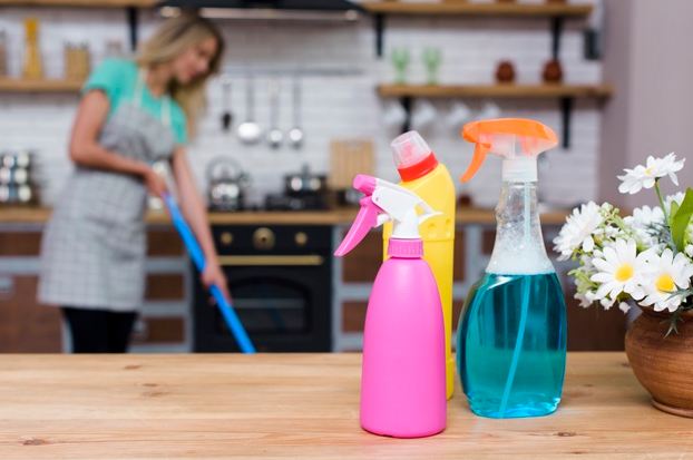 Image of detergents and spray bottles on a wooden desk with a woman mopping the floor in the background